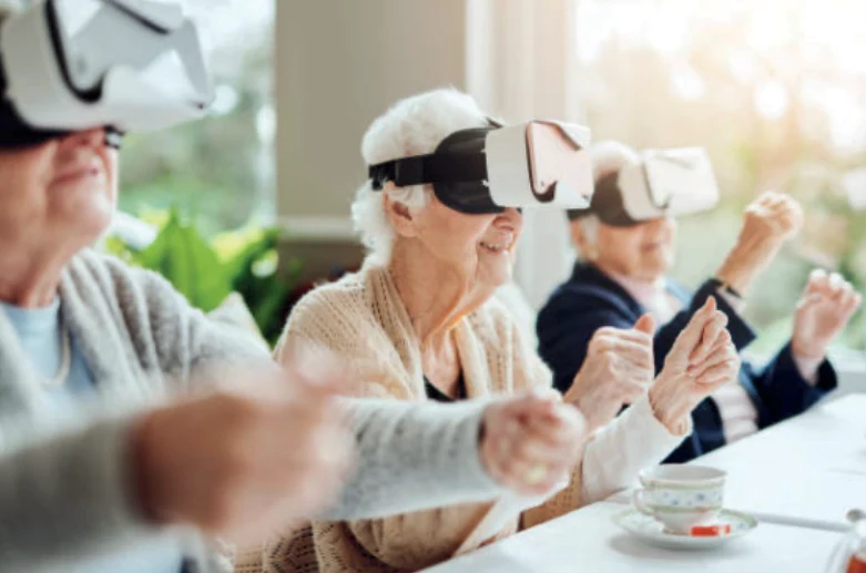 Personas mayores usando gafas de realidad virtual mientras realizan movimientos con las manos, sentadas en una mesa con luz natural.
