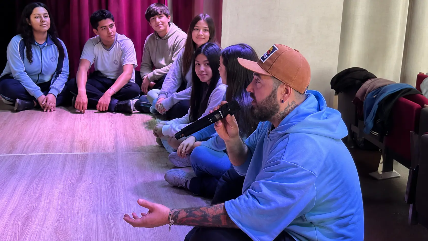Persona con un micrófono conversando con un grupo de jóvenes sentados en círculo durante una actividad.
