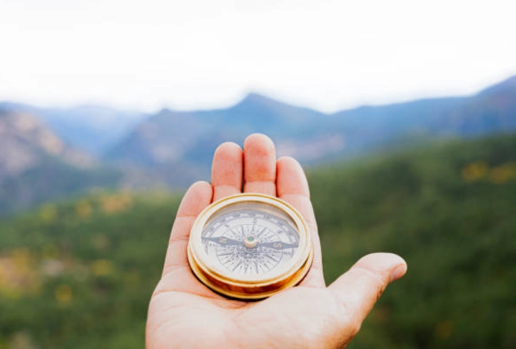 Mano sosteniendo una brújula dorada con paisaje montañoso desenfocado al fondo.