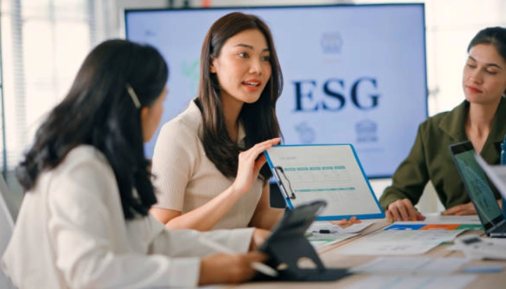 Tres mujeres reunidas en una sala de juntas; una de ellas sostiene una carpeta con documentos mientras habla, con una pantalla al fondo que muestra las letras ESG.
