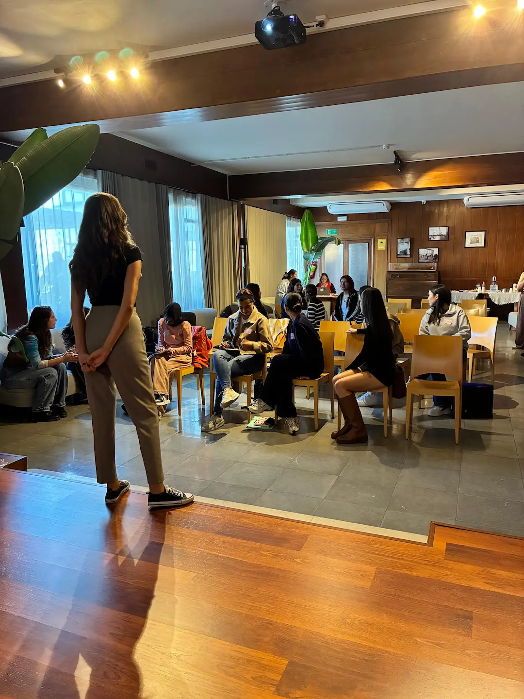 Grupo de personas sentadas en sillas en una sala interior, mientras una mujer está de pie al frente, en un entorno tipo taller o presentación.
