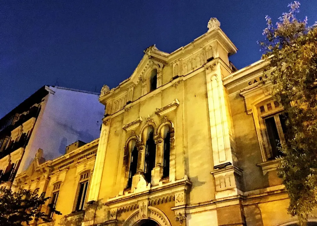 Edificio histórico iluminado por la noche, con fachada ornamentada y arquitectura clásica en una calle urbana.