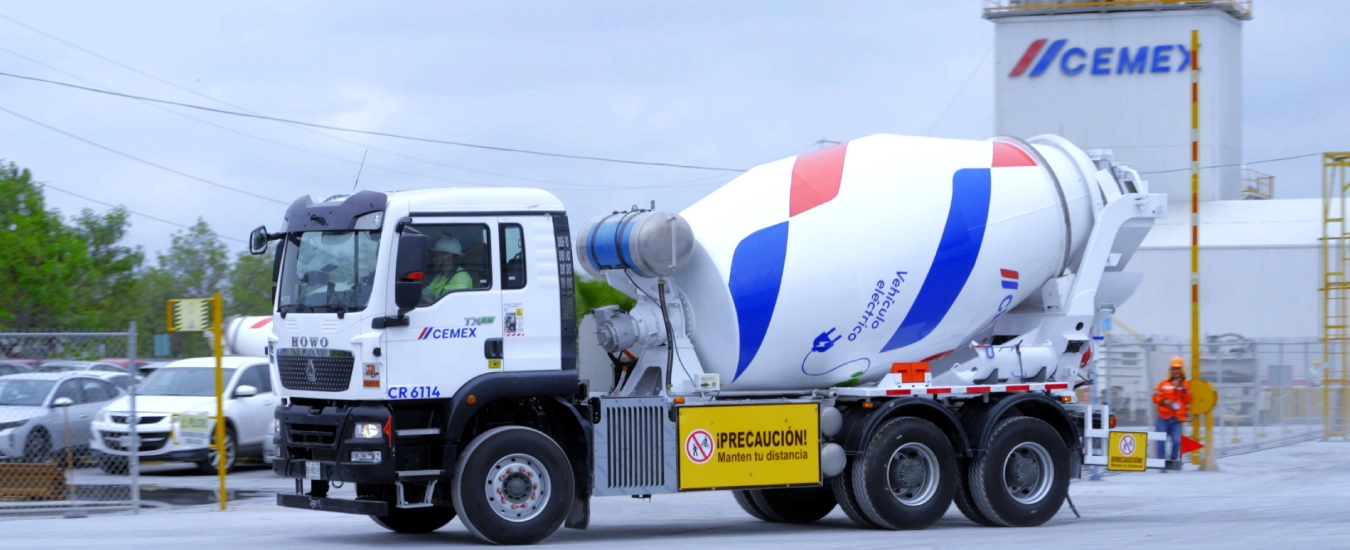 Camión mezclador de cemento de la empresa CEMEX en una planta industrial, con señalización de seguridad y vehículos al fondo.