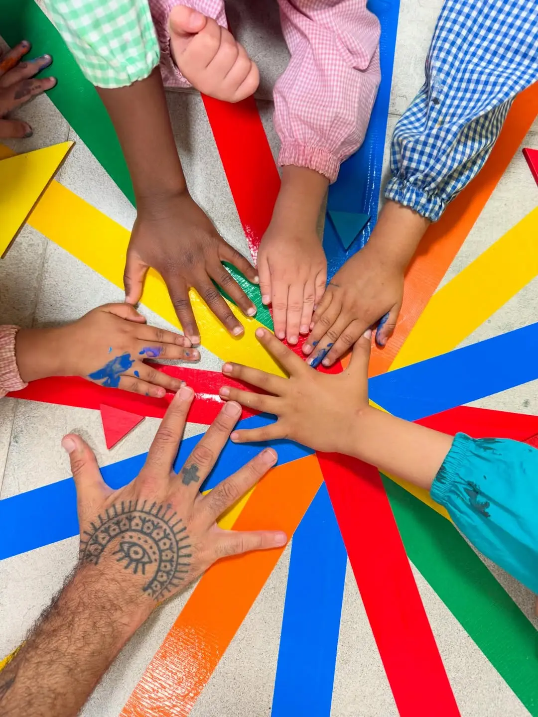 Manos de niños y un adulto unidas sobre una superficie con cintas de colores dispuestas en forma radial.