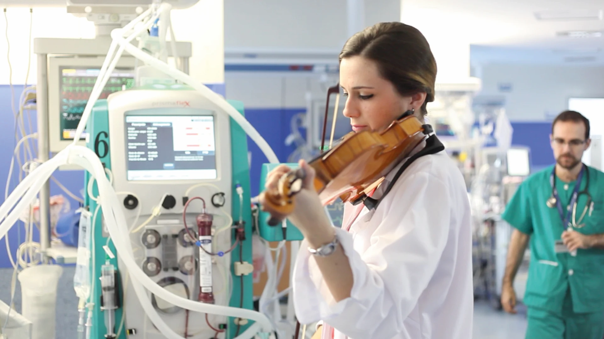 Mujer tocando un violín en una sala hospitalaria junto a equipos médicos, mientras un profesional de salud camina al fondo.