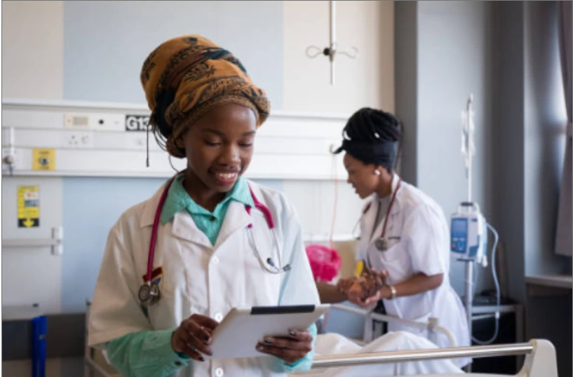 Profesional de la salud utilizando una tablet en una habitación hospitalaria, con otra enfermera atendiendo a un paciente en segundo plano.