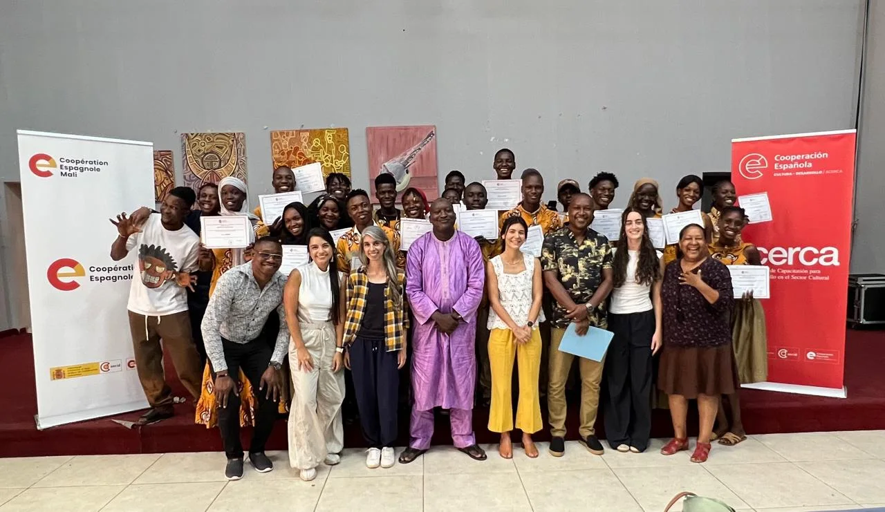 Grupo multicultural de personas posando con certificados en evento de cooperación internacional, frente a banners institucionales, destacando formación, colaboración y desarrollo comunitario.